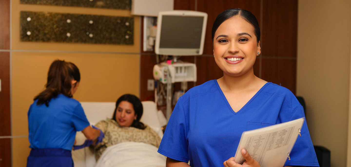 Smiling nurse in blue scrubs holding a clipboard, with a patient and another clinician in the background.