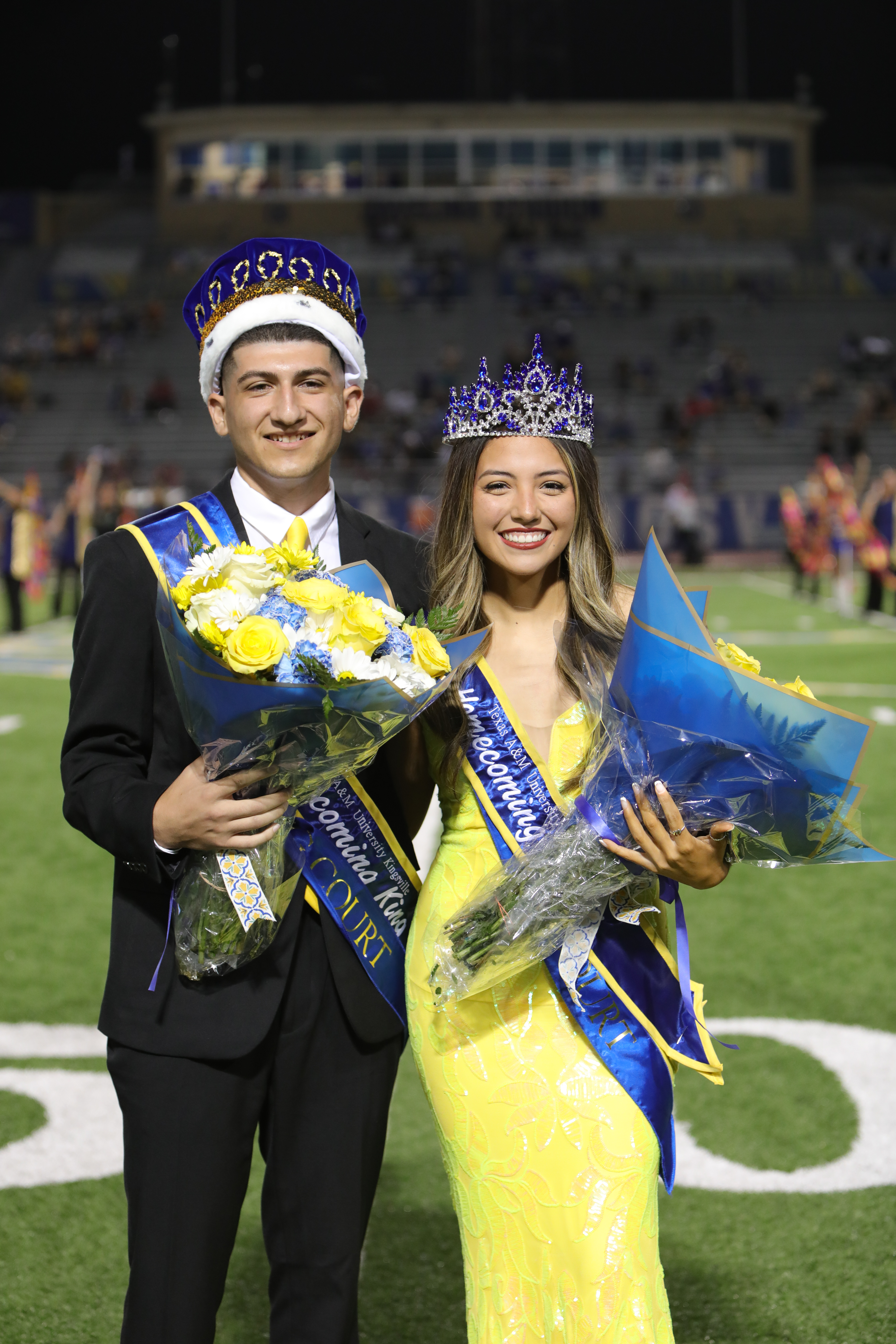 A king and queen pose together on the field, both holding bouquets of flowers and wearing crowns, during a homecoming celebration.