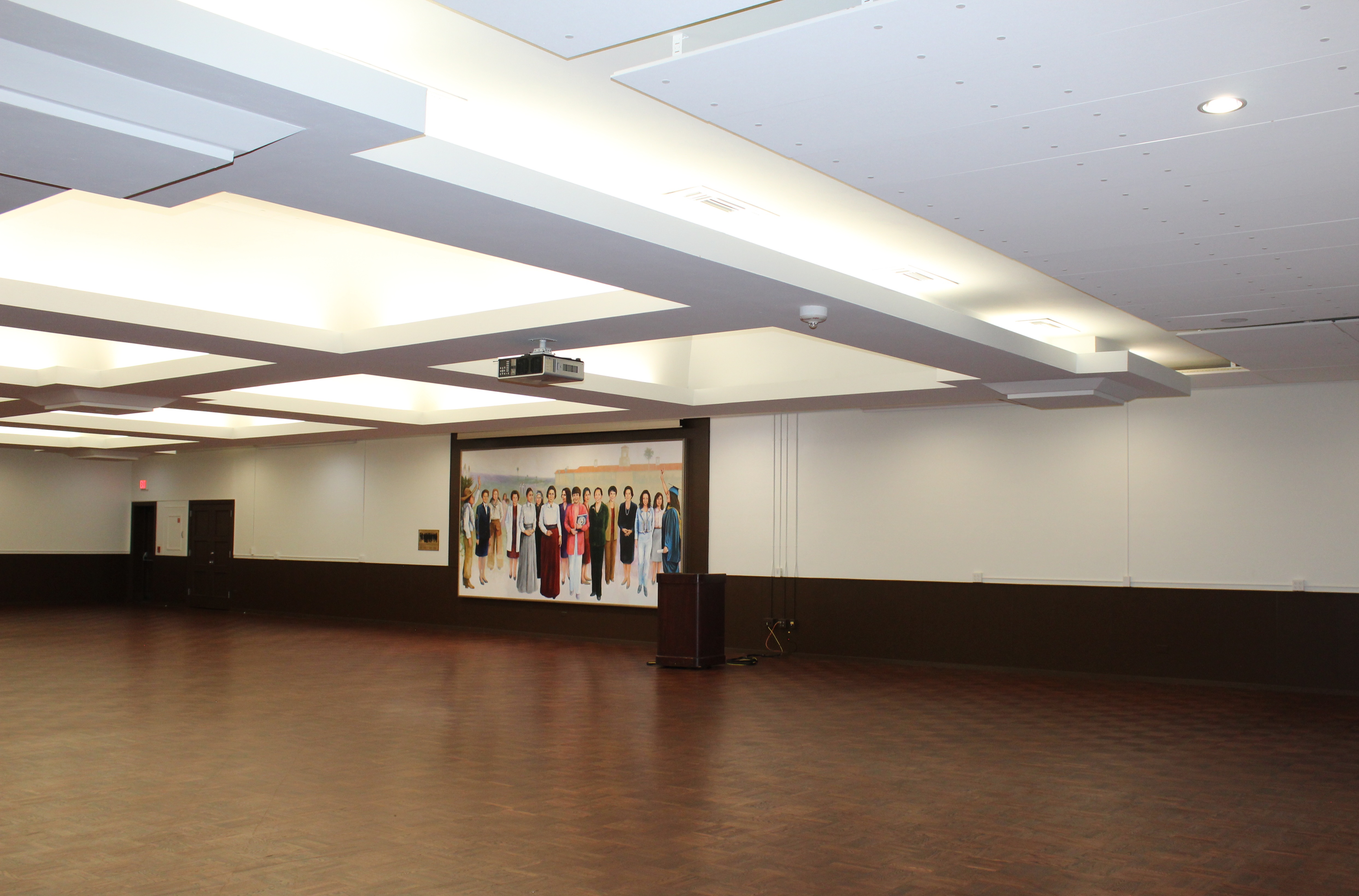 Ballroom A, empty, with a mural of important historical women of the university, and a room featuring lighted ceiling alcoves, newly painted white walls, and freshly stained flooring.