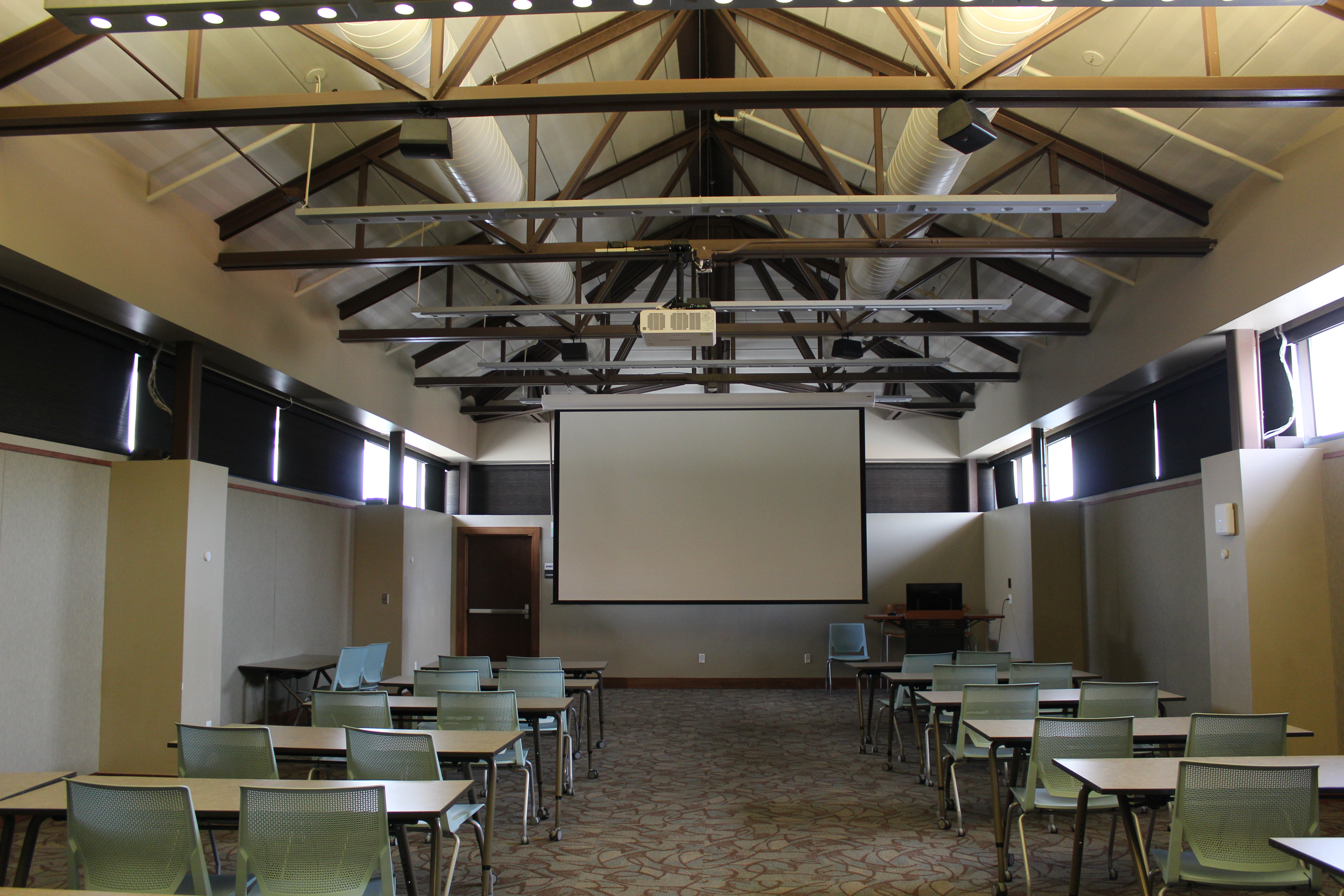 A large room with high ceilings and rafters, as well as a projector system. There are chairs and tables arranged to face the projector screen.