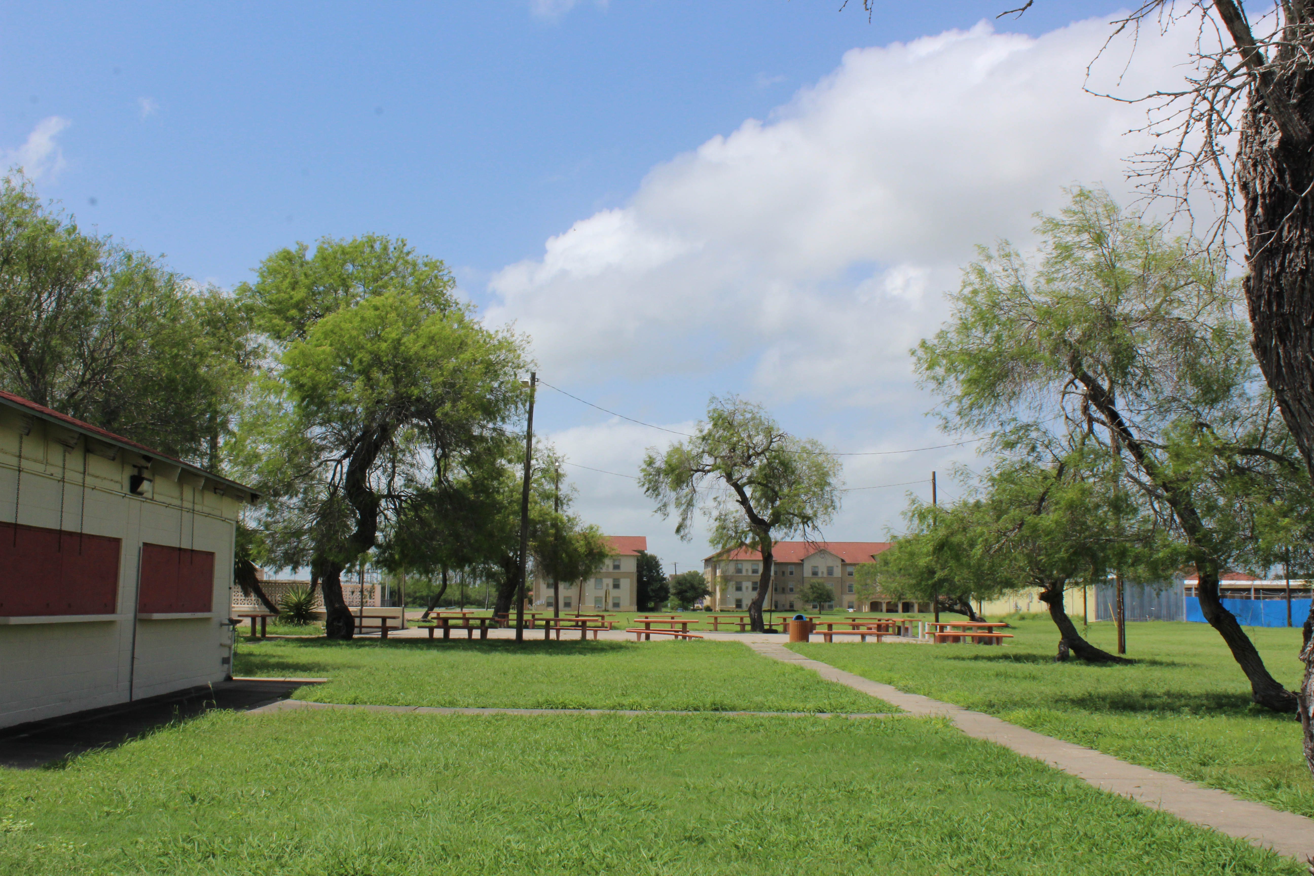 An image of a small building in front of a concrete courtyard ringed by bench tables. There is a large grassy area on all sides of the courtyard.