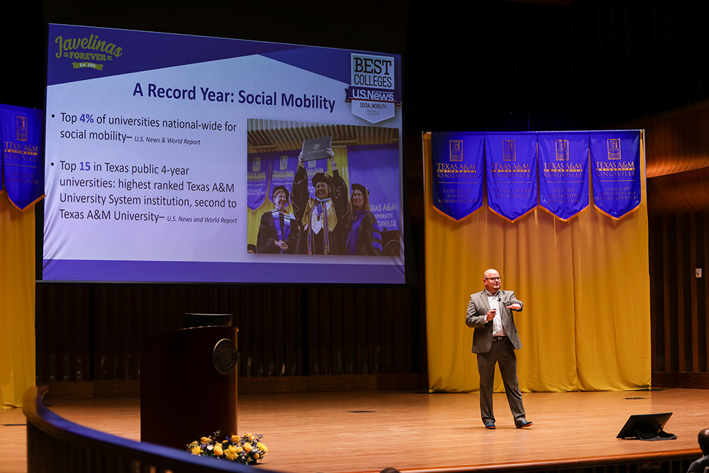 University president Dr. Robert Vela Jr. stands on the stage of the performance hall in the university's Music education complex.  Behind him is a PowerPoint presentation displaying a slide from his address presentation highlighting social mobility.