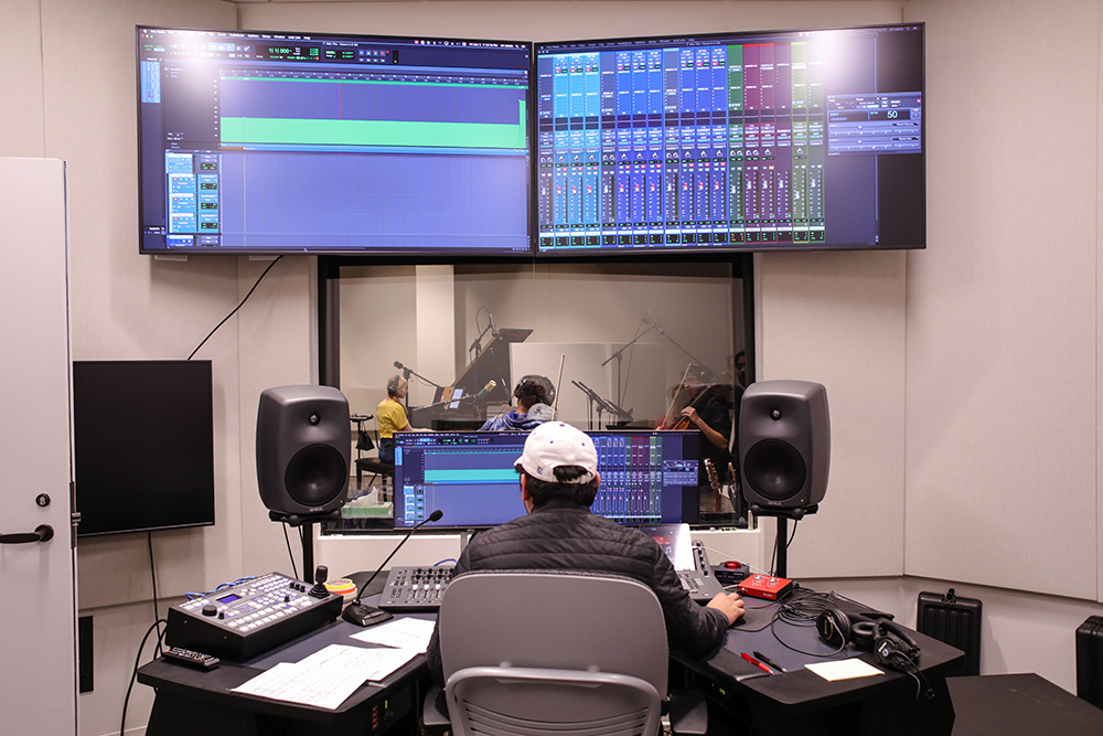 A Sound Recording Technology student sits before audio recording equipment and a large window as students play a variety of instruments in the next room.