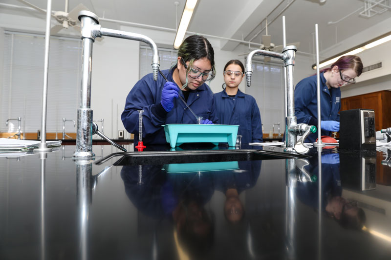 Female in blue chemistry lab coats puts liquid into a beaker using a dropper while another looks on in the background and another works off to the right side.
