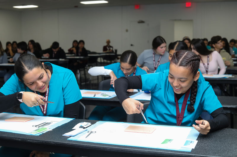 Two female students in green scrubs using scissors to cut thread to apply suture on lifelike skin pad.