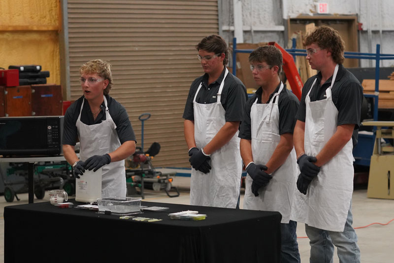 Four males in blue polo shirts and aprons give a presentation in an ag workshop