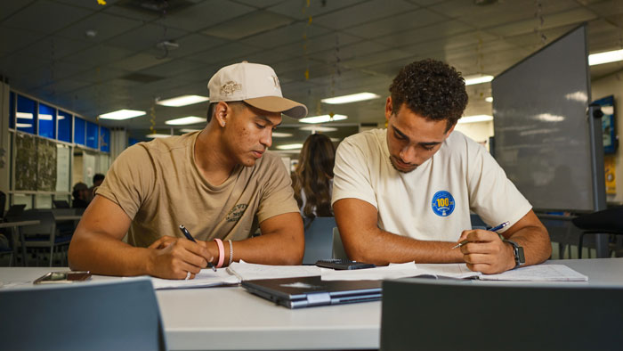 Two students sit at a table in a classroom, writing on paperwork together.