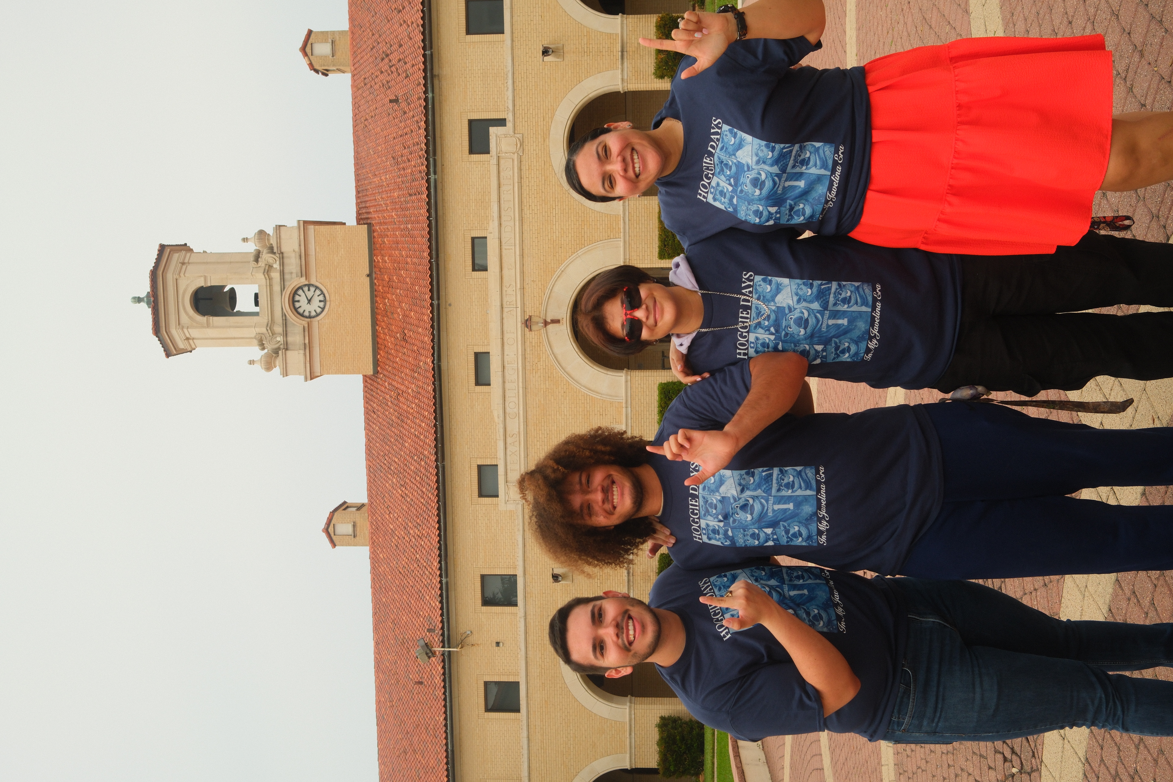 Four students in matching “Hoggie Days” shirts pose and smile in front of a campus building with a clock tower.